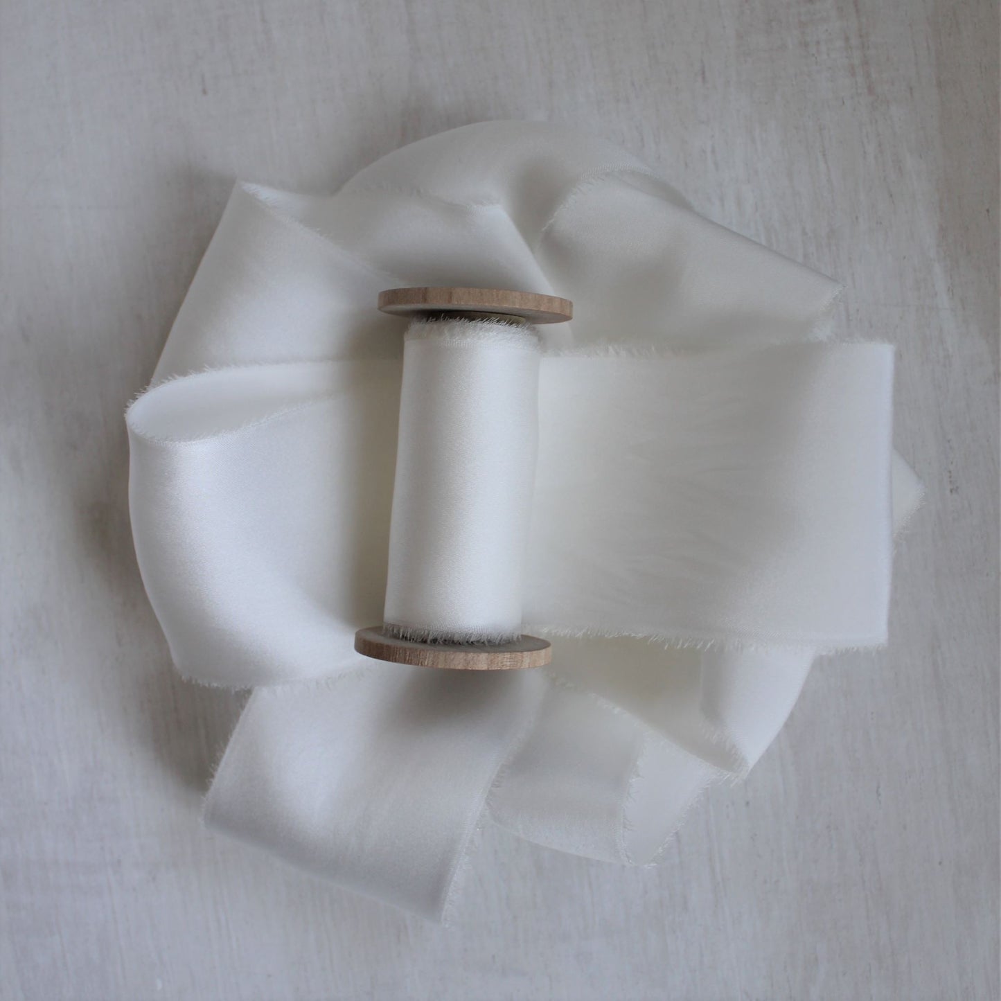 Hand-torn white silk charmeuse ribbon wound on a wooden spool, showcasing its soft sheen and frayed edges