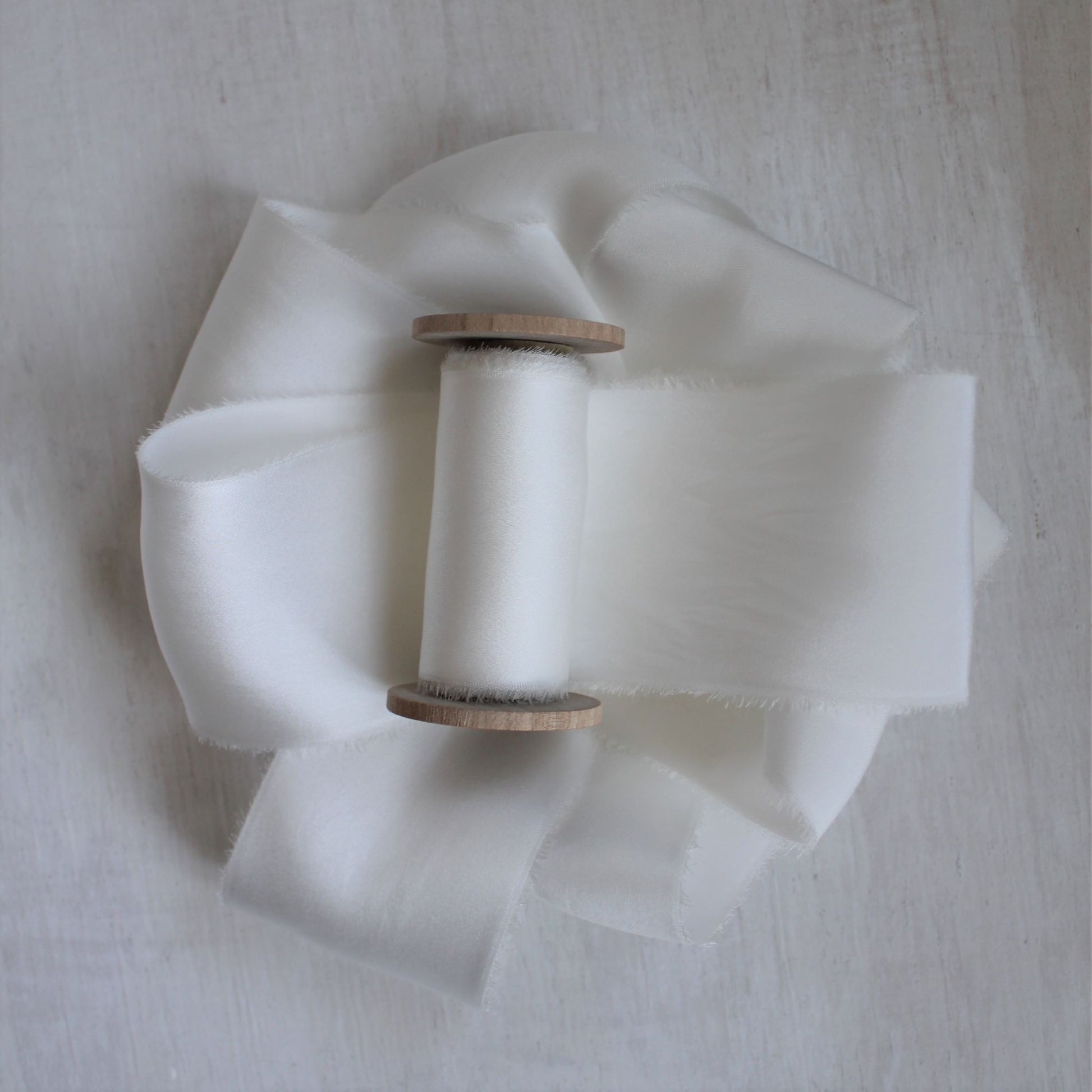 Hand-torn white silk charmeuse ribbon wound on a wooden spool, showcasing its soft sheen and frayed edges